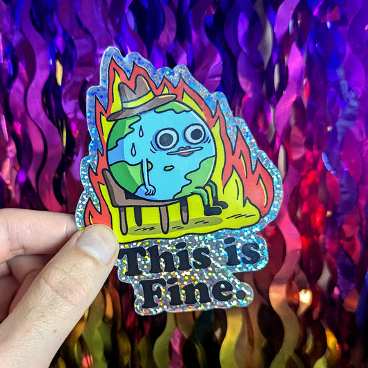 A vibrant, holographic-glitter vinyl sticker featuring a sweating cartoon Earth in flames, wearing a hat and the sarcastic text ‘This is Fine.’ Held against a rainbow sequin background, this waterproof, durable sticker is perfect for laptops, water bottles, and journals.