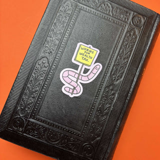 A close-up view of a black embossed notebook with a humorous sticker reading 'Worms for Wealth Tax' featuring a worm holding a sign, perfect for fans of quirky and unique stickers.