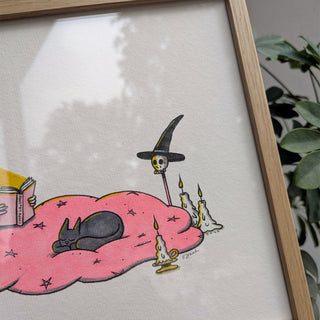 Framed illustration of a witch on a blanket with candles and a book.
