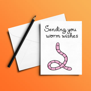 Funny worm greeting card with cute pink cartoon worm illustration and handwritten text 'Sending you worm wishes' on white card stock. Perfect for humorous birthday, encouragement, or get-well-soon messages. Blank inside for personalization. Shown with white envelope and black pencil on orange background.