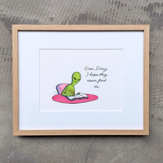 Framed artwork with an alien sitting on a pink planet, writing in a diary with text 'Dear Diary, I hope they never find us.'