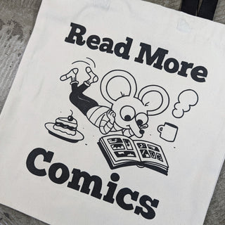 A white canvas tote bag with black printed text reading 'Read More Comics' and a cartoon mouse reading, photographed on a gray textured surface.
