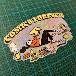 A die-cut glitter sticker featuring a cartoon character reading surrounded by books, with the text 'COMICS FOREVER' in yellow. The sticker has a holographic glitter background and is photographed on a green cutting mat.