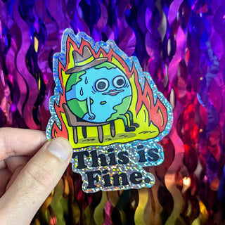 A vibrant, holographic-glitter vinyl sticker featuring a sweating cartoon Earth in flames, wearing a hat and the sarcastic text ‘This is Fine.’ Held against a rainbow sequin background, this waterproof, durable sticker is perfect for laptops, water bottles, and journals.
