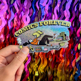 A hand holds a die-cut sticker featuring a comic book character lying on the floor surrounded by books, with the text 'COMICS FOREVER' arched above. The sticker has a holographic glitter background and is photographed in front of a colorful, metallic tinsel curtain.