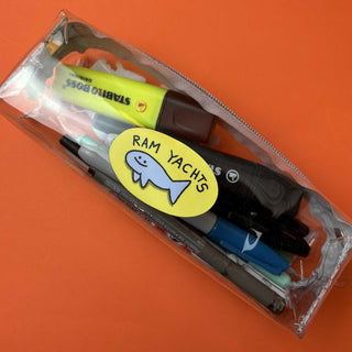 A transparent pencil case filled with pens, markers, and highlighters. A yellow “RAM YACHTS” sticker with a cartoon fish is stuck on the outside of the case.