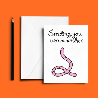 Whimsical worm-themed greeting card with adorable pink worm drawing and handwritten 'Sending you worm wishes' text. Great for unique, funny, or cheerful greetings. Blank interior for your personal message. Shown with white envelope and pencil on vibrant orange background.